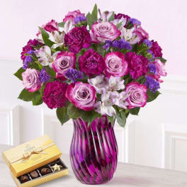 Pink and purple bouquet in a purple glass vase with a box of chocolates