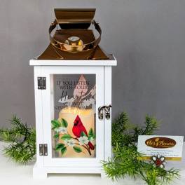 White lantern with a lit candle and cardinal design, surrounded by greenery.