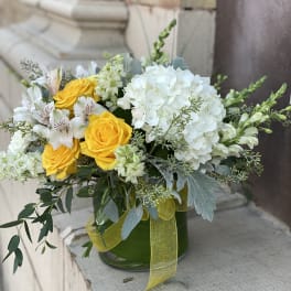 Low glass vase of yellow roses, white hydrangeas, and white blooms with a yellow ribbon.