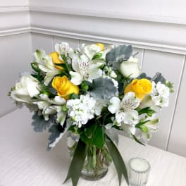 Yellow and white roses with white alstroemeria and soft gray foliage in a clear glass vase