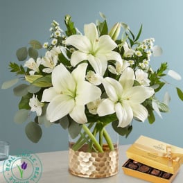 White lilies in a glass vase with a box of chocolates beside it