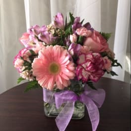 Compact pink and purple flower arrangement in a clear glass cube vase with a lavender ribbon bow