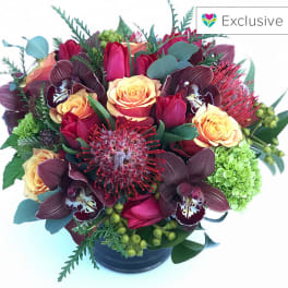 Low round arrangement of red and peach flowers with orchids and pincushion protea in a dark vase