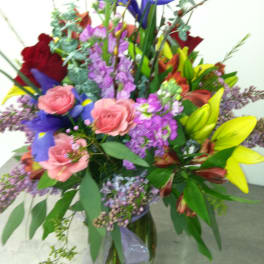 Mixed bouquet of pink roses, purple blooms, yellow lilies, and red flowers in a clear glass vase with ribbon