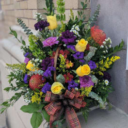 Basket arrangement with yellow roses, purple blooms, and a plaid ribbon