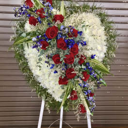 Tall heart-shaped standing spray with white flowers, red roses, and blue blooms on a white easel.