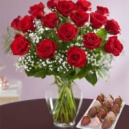 Red roses in a glass vase beside a tray of chocolate-covered strawberries