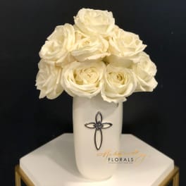 White roses arranged in a tall white vase with a cross emblem