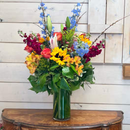Tall mixed arrangement with sunflowers, roses, lilies, and blue delphinium in a clear glass cylinder vase.