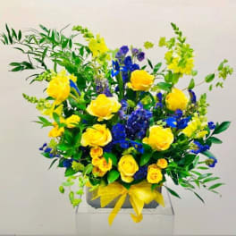 Yellow roses and blue flowers arranged in a square vase with a yellow ribbon.