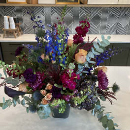 Large mixed floral arrangement in a dark vase with blue, purple, and peach blooms