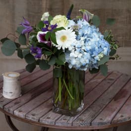 Bouquet of blue hydrangeas, white gerbera daisy, and purple blooms in a glass vase