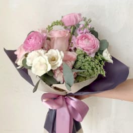 Hand-tied bouquet of pink and white flowers wrapped in dark paper with a satin ribbon.
