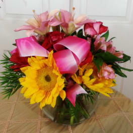 Bouquet of sunflowers, pink lilies, and roses with pink ribbon in a glass vase