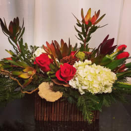 Low floral arrangement with red roses, white hydrangea, and tropical blooms in a rustic container