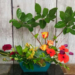 Mixed floral arrangement in a teal container with bright blooms and tall leafy branches