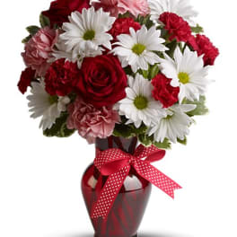 Red roses, white daisies, and pink carnations in a red vase with a ribbon