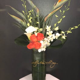 Tropical bouquet with orange roses, white calla lilies, and orchids in a tall glass vase