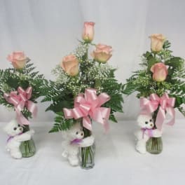 Three pink rose arrangements in glass vases with teddy bears and ribbons