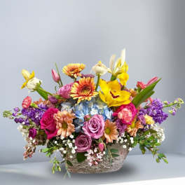 Colorful arrangement of roses, gerbera daisies, orchids, and mixed blooms in a low ceramic container
