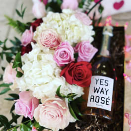 Pink and white roses with hydrangeas beside a bottle of rosé in a gift box