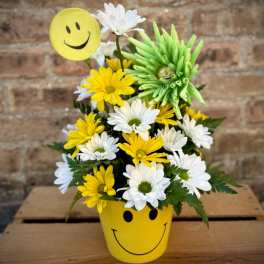 Yellow smiley-face pot holding white and yellow daisies with a smiley pick