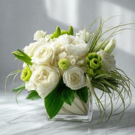 Low white rose and carnation arrangement with green ranunculus in a square glass vase and arching grass accents.
