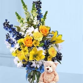 Tall yellow, white, and blue flower arrangement in a vase with a tan teddy bear