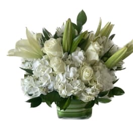Low white arrangement of roses, lilies, and hydrangeas in a clear glass vase with green lining