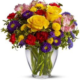 Mixed bouquet of yellow roses, red carnations, and purple daisies in a glass vase