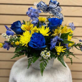 Blue roses and yellow daisies in a white vase with a patterned ribbon bow