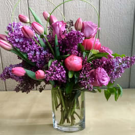 Pink tulips, roses, peonies, and purple lilacs arranged in a clear glass vase