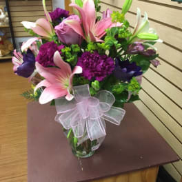 Pink lilies and purple flowers arranged in a glass vase with a sheer ribbon