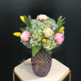Mixed bouquet in a purple glass vase with blue hydrangeas, pink peonies, and yellow tulips