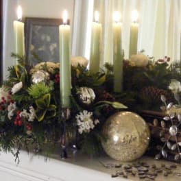 Holiday candle centerpiece with greenery and ornaments on a mantel