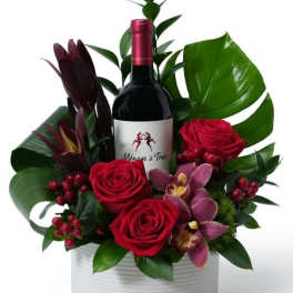 Red wine gift arrangement with red roses, pink orchids, berries, and large green leaves in a white round container