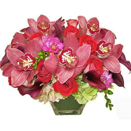 Low arrangement of pink cymbidium orchids, red roses, and dark calla lilies in a clear glass vase