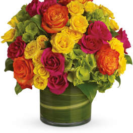 Compact arrangement of yellow, orange, and pink roses with green hydrangeas in a green glass cylinder vase