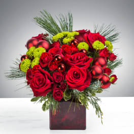 Red rose arrangement with green button mums, pine accents, and red ornaments in a red glass cube vase.