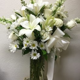 White lilies and daisies arranged in a tall glass vase with a white ribbon.
