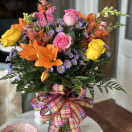 Colorful bouquet of roses, lilies, and purple daisies in a glass vase with a plaid ribbon.