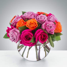 Short arrangement of hot pink, lavender, and orange roses in a round clear glass vase
