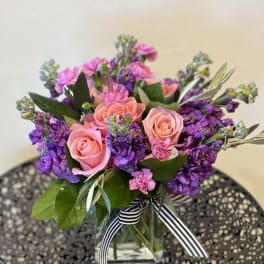 Pink roses and purple stock in a clear glass vase tied with a black and white striped ribbon.