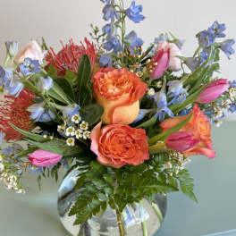 Bouquet of orange roses, pink tulips, and blue flowers in a glass vase