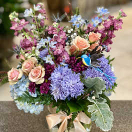 Mixed bouquet of pastel roses, purple blooms, and blue flowers in a glass vase