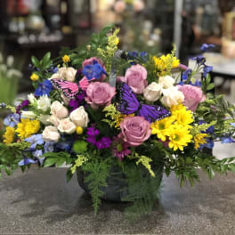Mixed bouquet of pink roses, yellow daisies, and blue flowers in a black basket