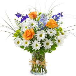 Mixed bouquet of white daisies, orange roses, and purple flowers in a clear vase tied with raffia and a shell.
