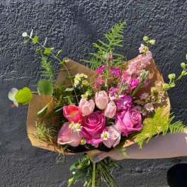 Hand-tied bouquet of pink roses, tulips, and small white daisies wrapped in brown paper.
