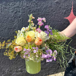 Pastel arrangement of yellow roses, peach tulips, and purple blooms in a light green vase.