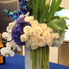 Tall floral arrangement with white roses, orchids, and blue-purple flowers in a white vase